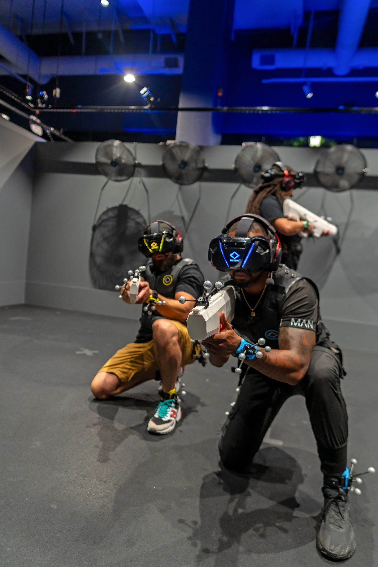 Players in VR headsets and vests crouching mid game under blue arena light